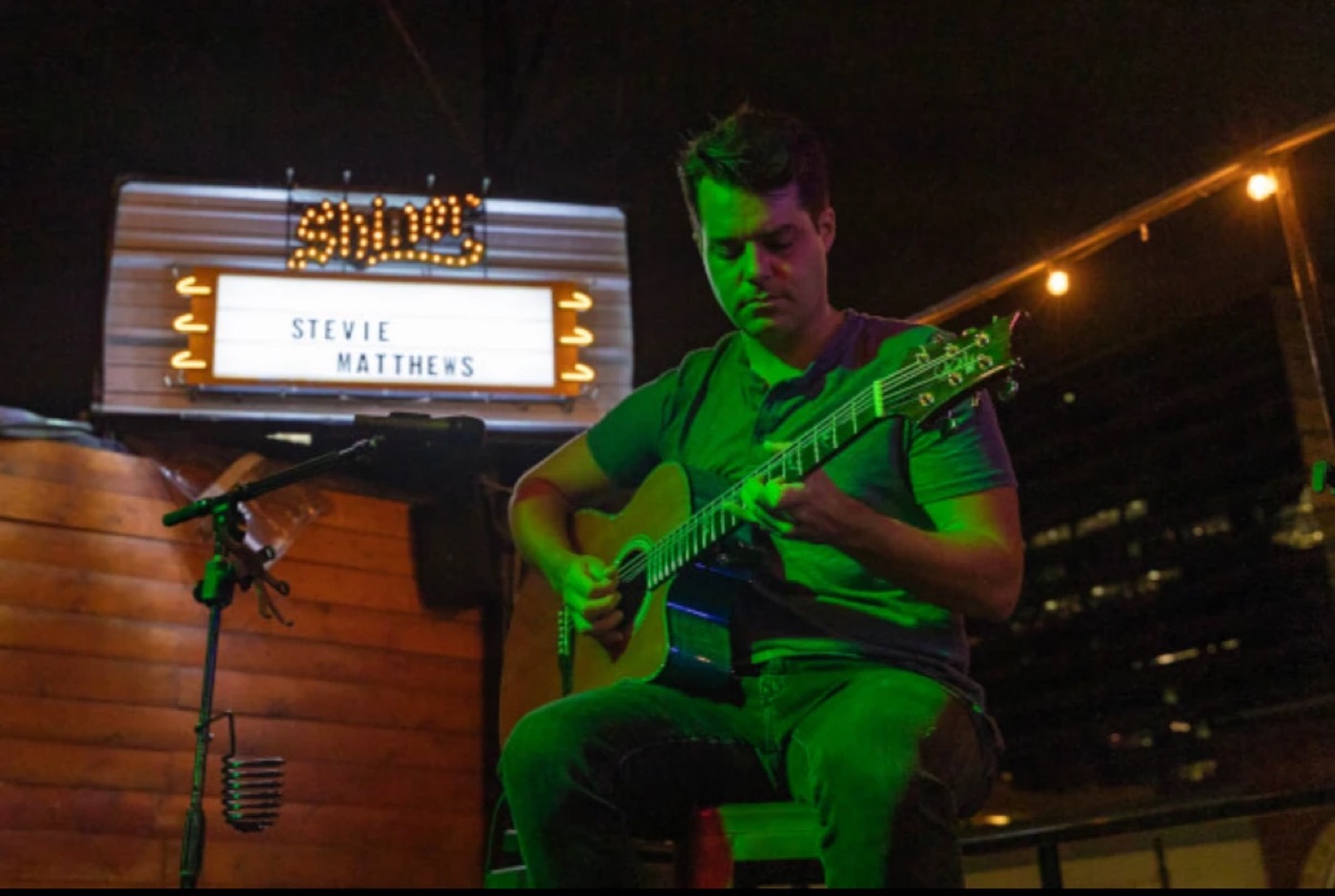 Stevie Matthews playing acoustic guitar under a lit venue marquee showing his name
